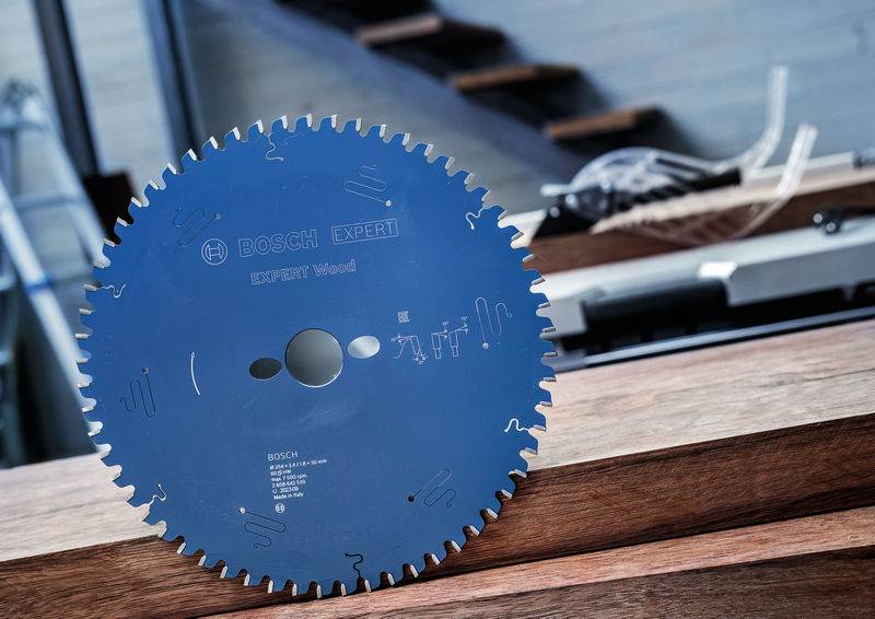 Ein blaues kreisförmiges Sägeblatt mit der Aufschrift „Bosch Expert Wood" ist aufrecht auf Holzbrettern gestützt, mit Treppen im Hintergrund.