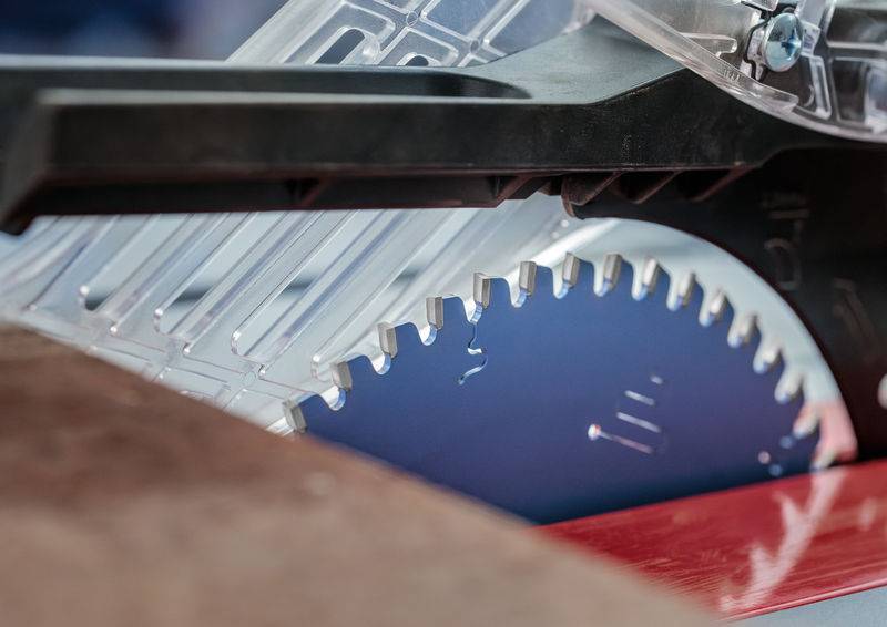 Eine Nahaufnahme einer Kreissägeblatt, das teilweise von einer transparenten Schutzvorrichtung bedeckt ist, zeigt scharfe Metallzähne auf blauem Hintergrund.