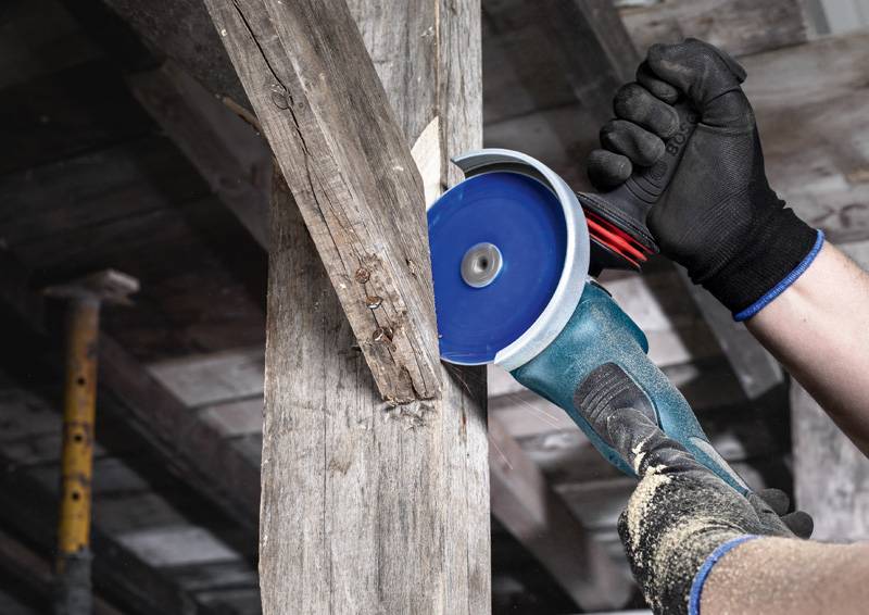 Person, die einen Winkelschleifer auf einem Holzbalken in einer Werkstattumgebung benutzt und dabei Schutzhandschuhe trägt.