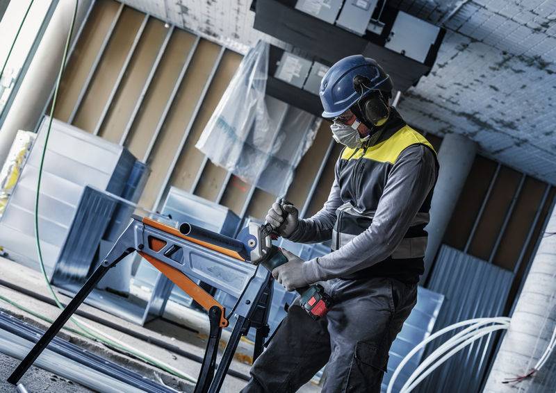 Ein Bauarbeiter in Schutzausrüstung bedient eine Säge auf einer Werkbank in einem unfertigen Industriegebäude.