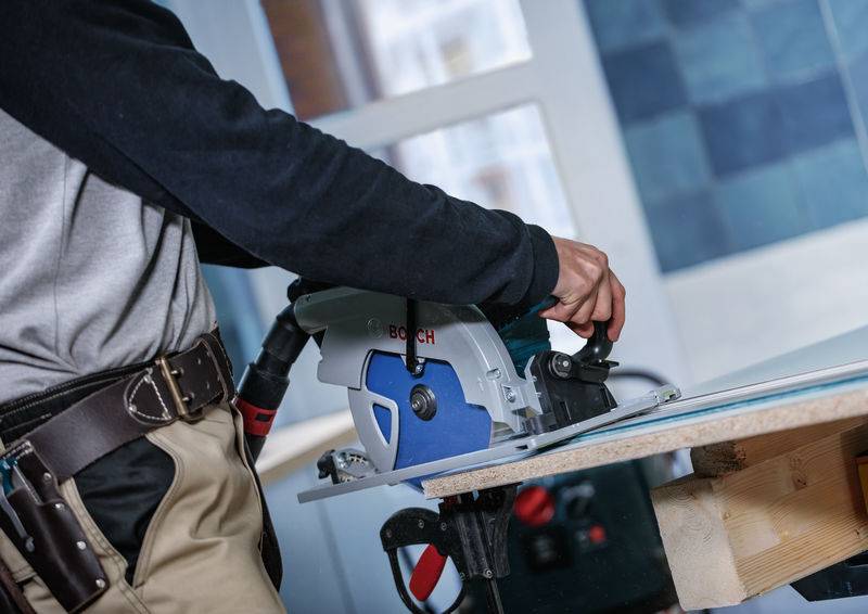 Eine Person benutzt eine Kreissäge, um ein Holzbrett in einer Werkstatt zu schneiden. Sie trägt einen Werkzeuggürtel, was auf handwerkliche Geschicklichkeit oder Zimmererarbeit hindeutet.