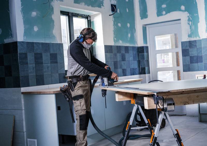 Eine Person in Schutzausrüstung bedient einen Elektroschleifen auf einer Holzplatte in einem Renovierungsraum mit teilweise fertiggestellten Wänden.