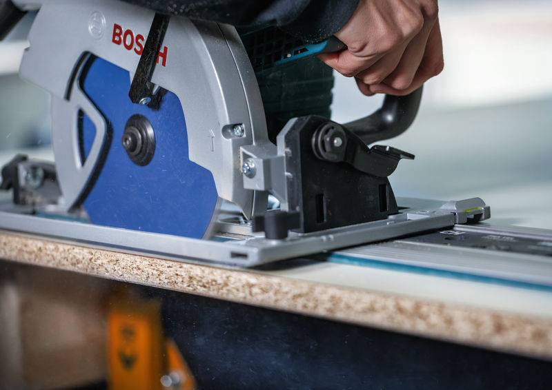 Eine Person bedient eine Kreissäge, um ein Holzstück auf einer Werkbank zu schneiden, wobei ein blaues Sägeblatt sichtbar ist.