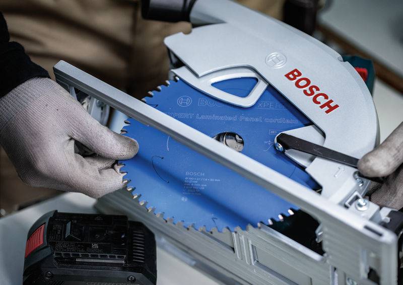 Eine Person in Handschuhen stellt ein blaues kreisförmiges Sägeblatt an einem Bosch-Elektrowerkzeug ein, wobei die Wartung oder Einrichtung von Holzbearbeitungsgeräten hervorgehoben wird.