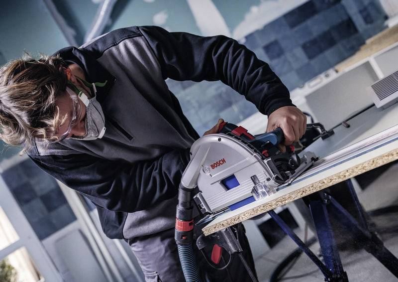 Eine Person in Schutzausrüstung bedient eine Bosch-Elektrosäge auf einer Sperrholzplatte in einer Werkstattumgebung und konzentriert sich auf präzises Schneiden.