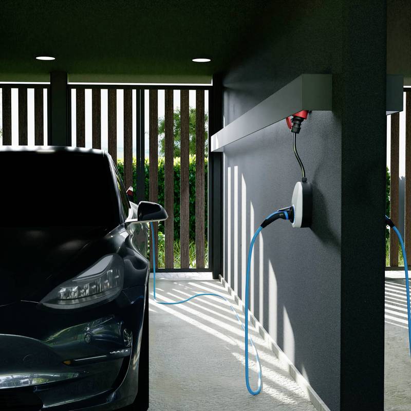 Ein Elektroauto ist in einer Garage geparkt und mit einer Ladestation verbunden. Sonnenlicht erzeugt gestreifte Schatten an der Wand, die auf eine teilweise Umschließung hindeuten.