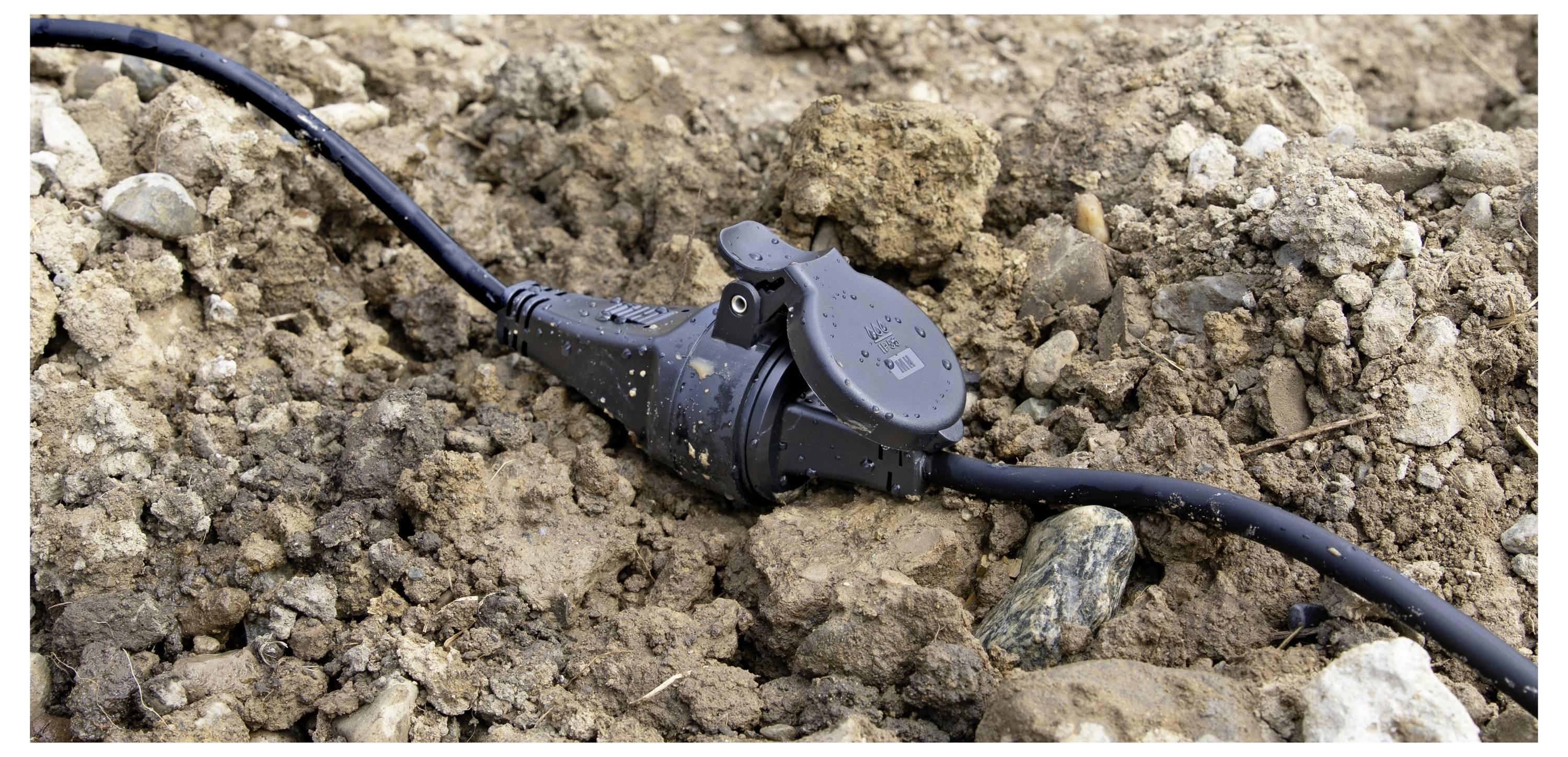 Ein wasserdichter Elektrostecker liegt teilweise im Boden vergraben, umgeben von Steinen und Erde, was auf die Verwendung im Freien hinweist.