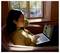 Eine Frau sitzt in einem Sessel und arbeitet am Laptop. Sonnenlicht fällt durchs Fenster. Draußen sind herbstliche Bäume zu sehen.