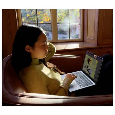 Eine Person sitzt in einem Stuhl vor einem Fenster und arbeitet an einem Laptop. Draußen sind Herbstbäume sichtbar.