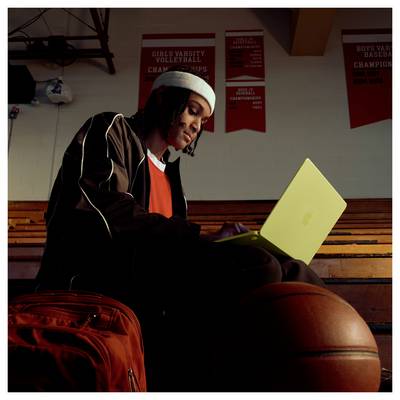 Eine Person sitzt in einer Sporthalle auf Zuschauertribünen mit einem gelben Laptop, einem Basketball und einem Rucksack. Banner zeigen Volleyball- und Basketballmeisterschaften.