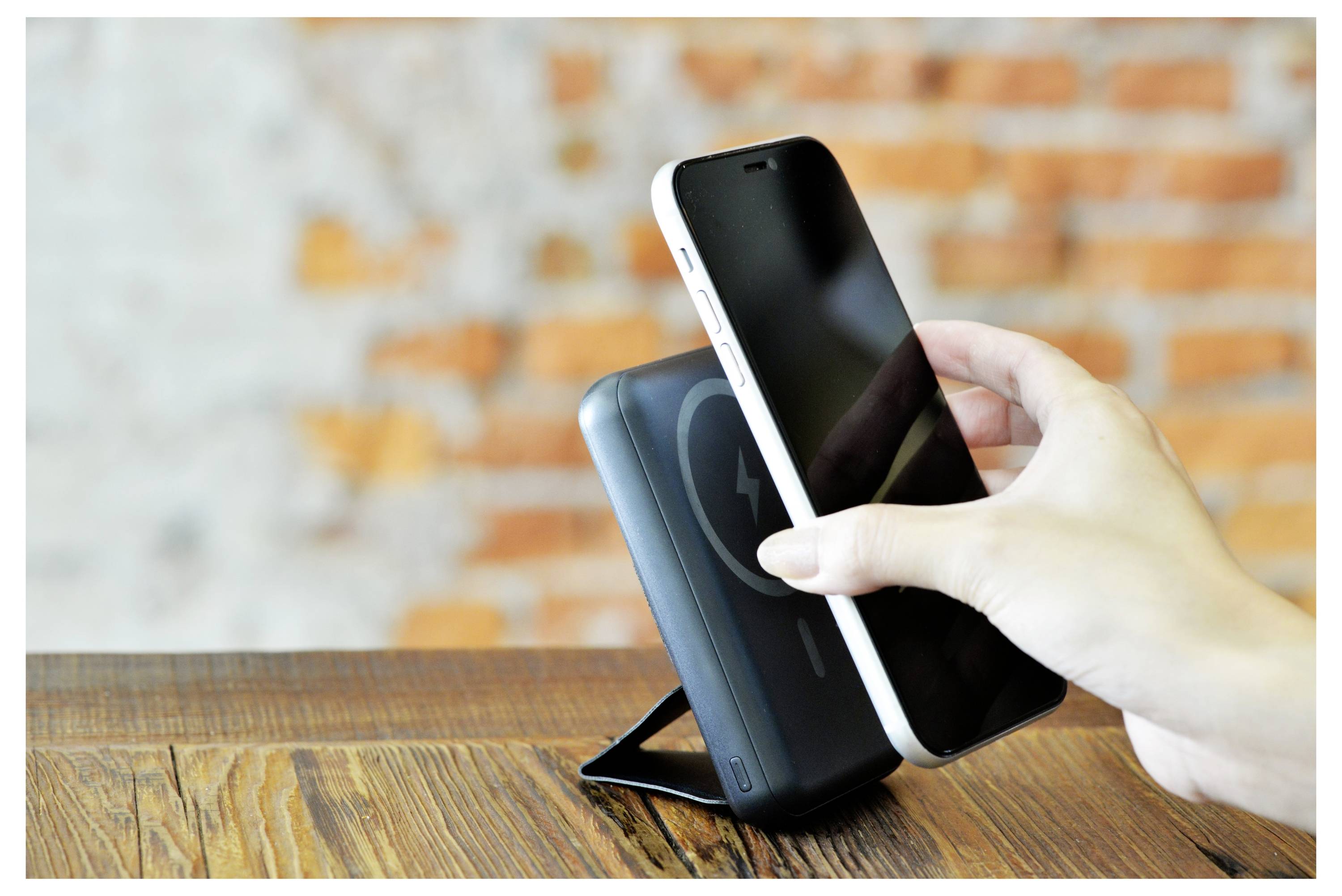 Eine Hand hält ein Smartphone über eine kabellose Ladestation auf einer Holzoberfläche, mit einer Backsteinmauer im Hintergrund.