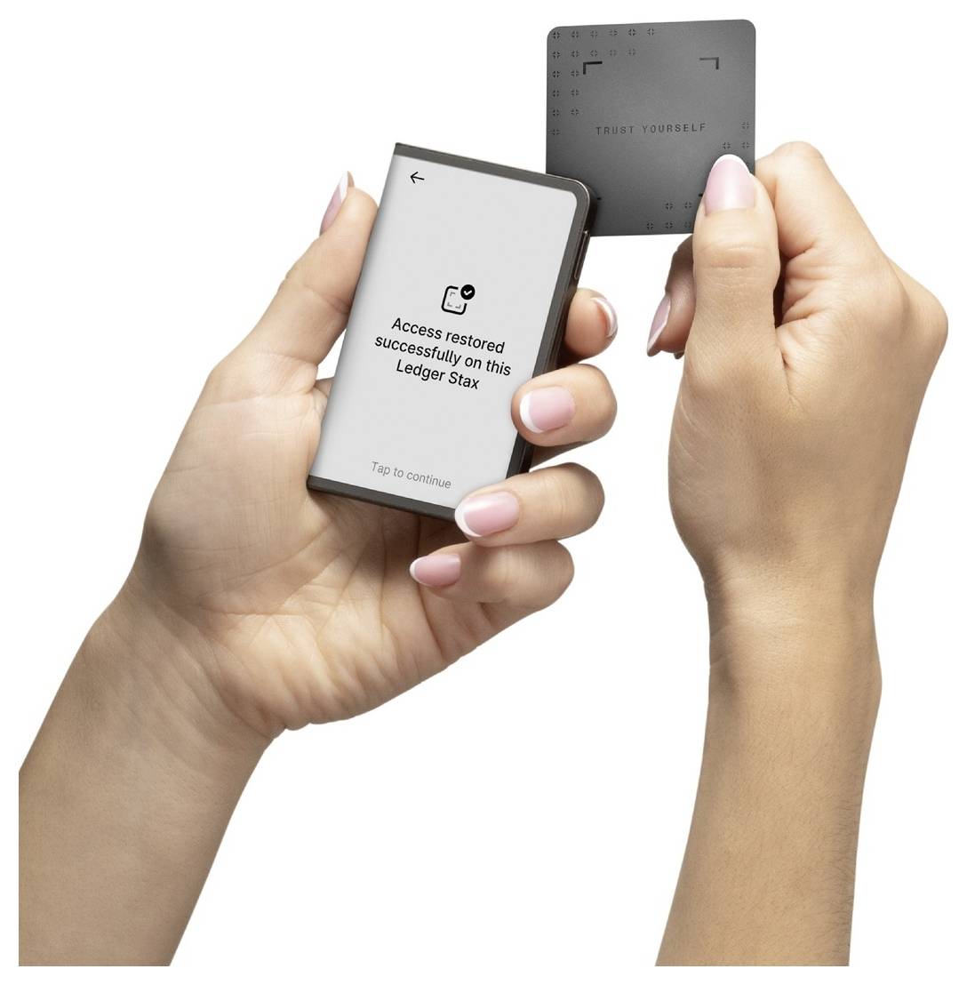 Zwei Hände halten ein Gerät und eine Karte. Der Bildschirm zeigt: „Zugriff auf diesem Ledger Stax erfolgreich wiederhergestellt. Zum Fortfahren tippen."