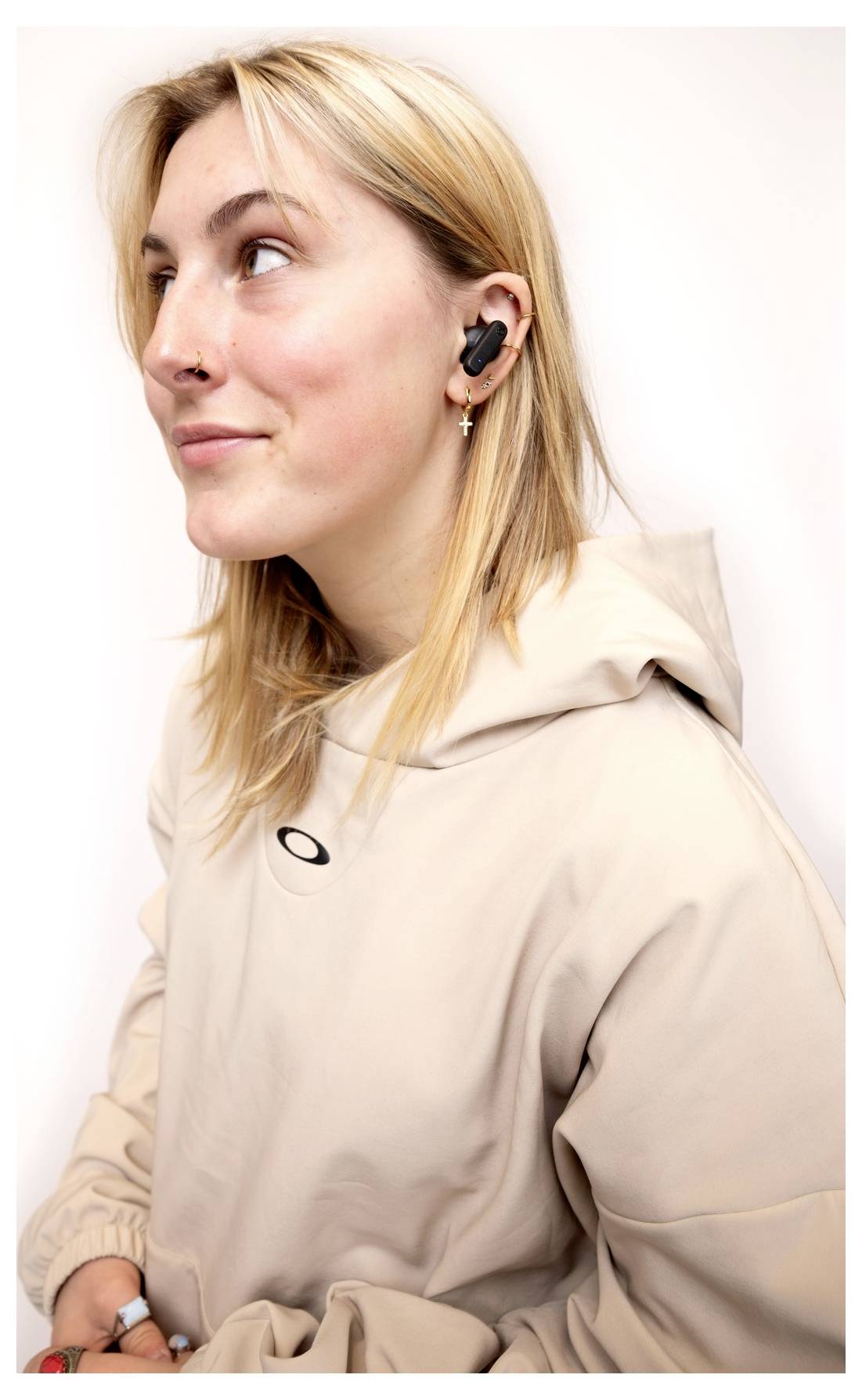 Eine Person, die drahtlose In-Ear-Kopfhörer und einen hellbeigen Hoodie trägt, blickt gegen einen weißen Hintergrund nach oben.