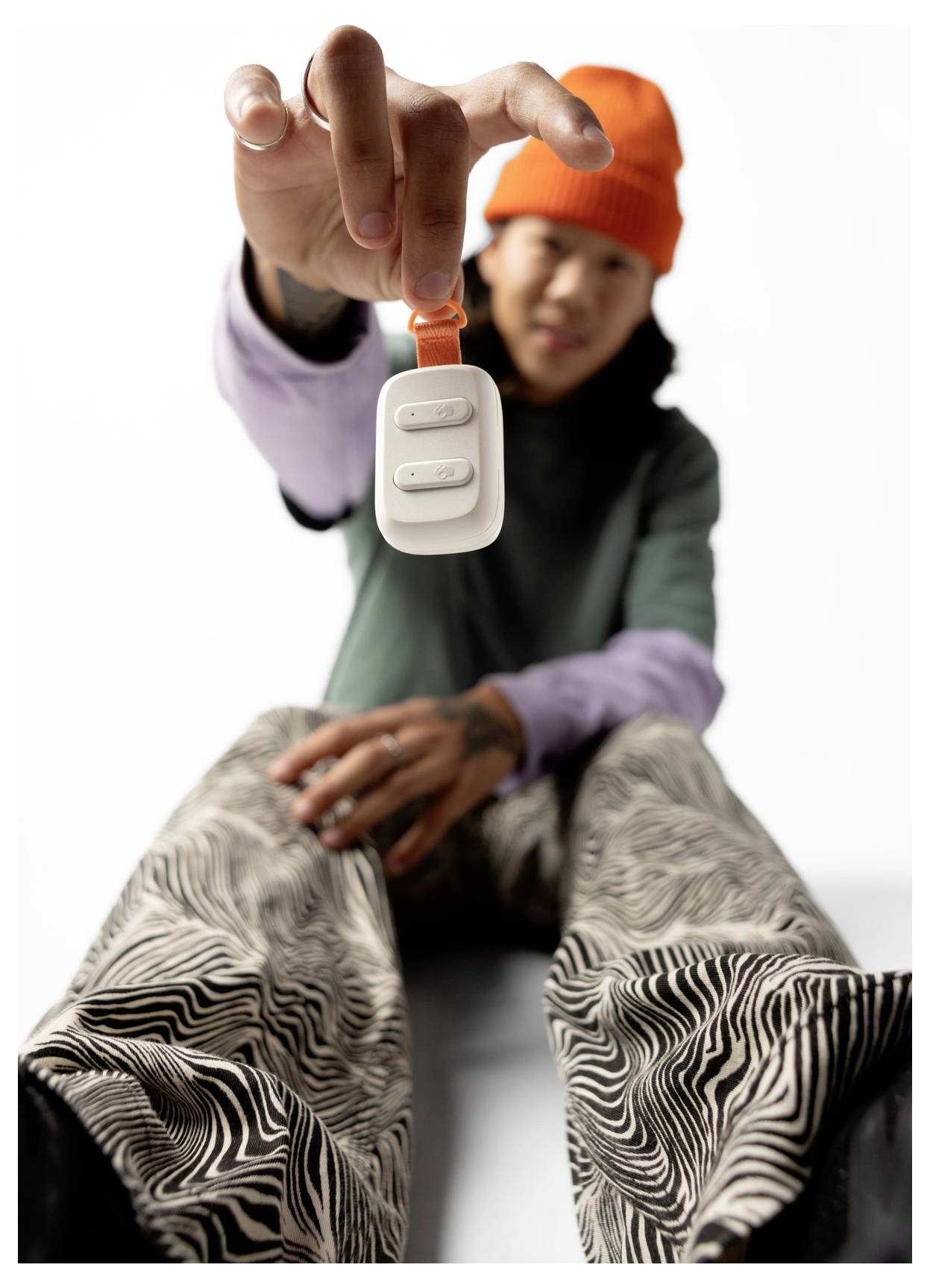 Eine Person sitzt und streckt die Hand aus, die ein kleines, weißes Gerät mit Tasten hält. Sie trägt eine leuchtend orangefarbene Beanie und gemusterte Hosen.