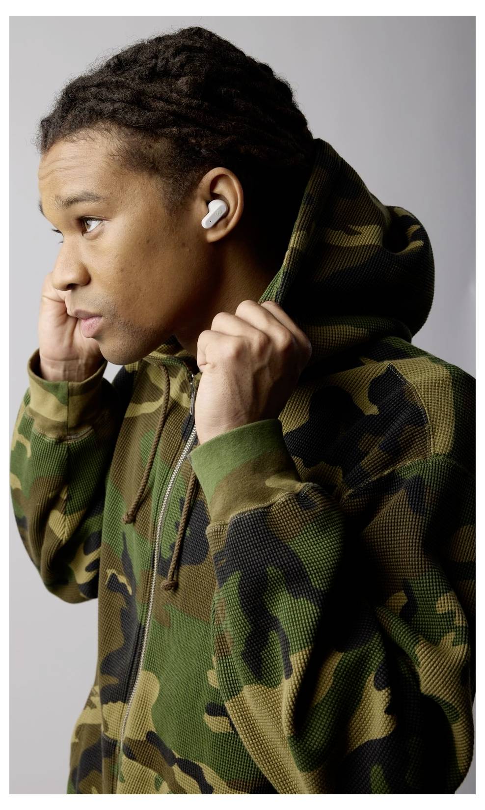Eine Person mit einem Camouflage-Hoodie und kabellosen Ohrhörern, die die Kapuze während einer Seitenwende justiert, vor einem einfachen Hintergrund.