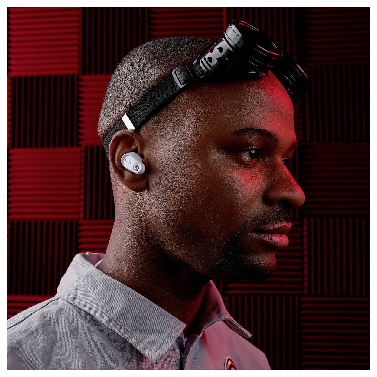 Eine Person in einem grauen Hemd, mit einem Ohrhörer und schwarzen Schutzbrille auf dem Kopf, steht vor einem rot-schwarz karierten Hintergrund.