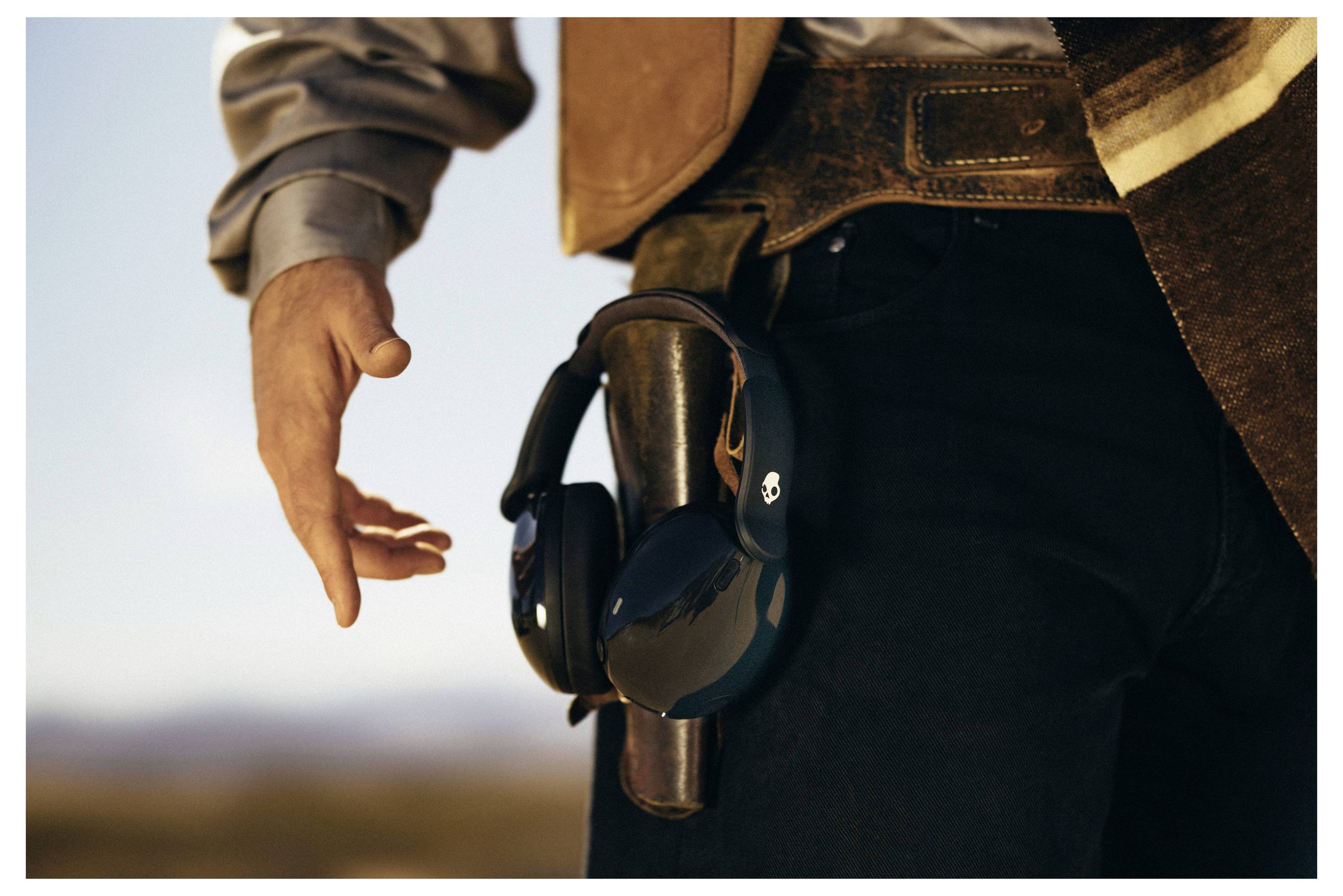 Eine Person, die eine Jeans und eine Jacke trägt, greift nach einem Paar schwarzer Kopfhörer, die an ihrem Gürtel befestigt sind, vor dem Hintergrund einer unscharfen Außenlandschaft.