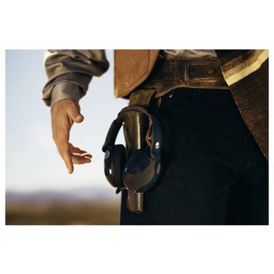 Eine Person, die eine Jeans und eine Jacke trägt, greift nach einem Paar schwarzer Kopfhörer, die an ihrem Gürtel befestigt sind, vor dem Hintergrund einer unscharfen Außenlandschaft.