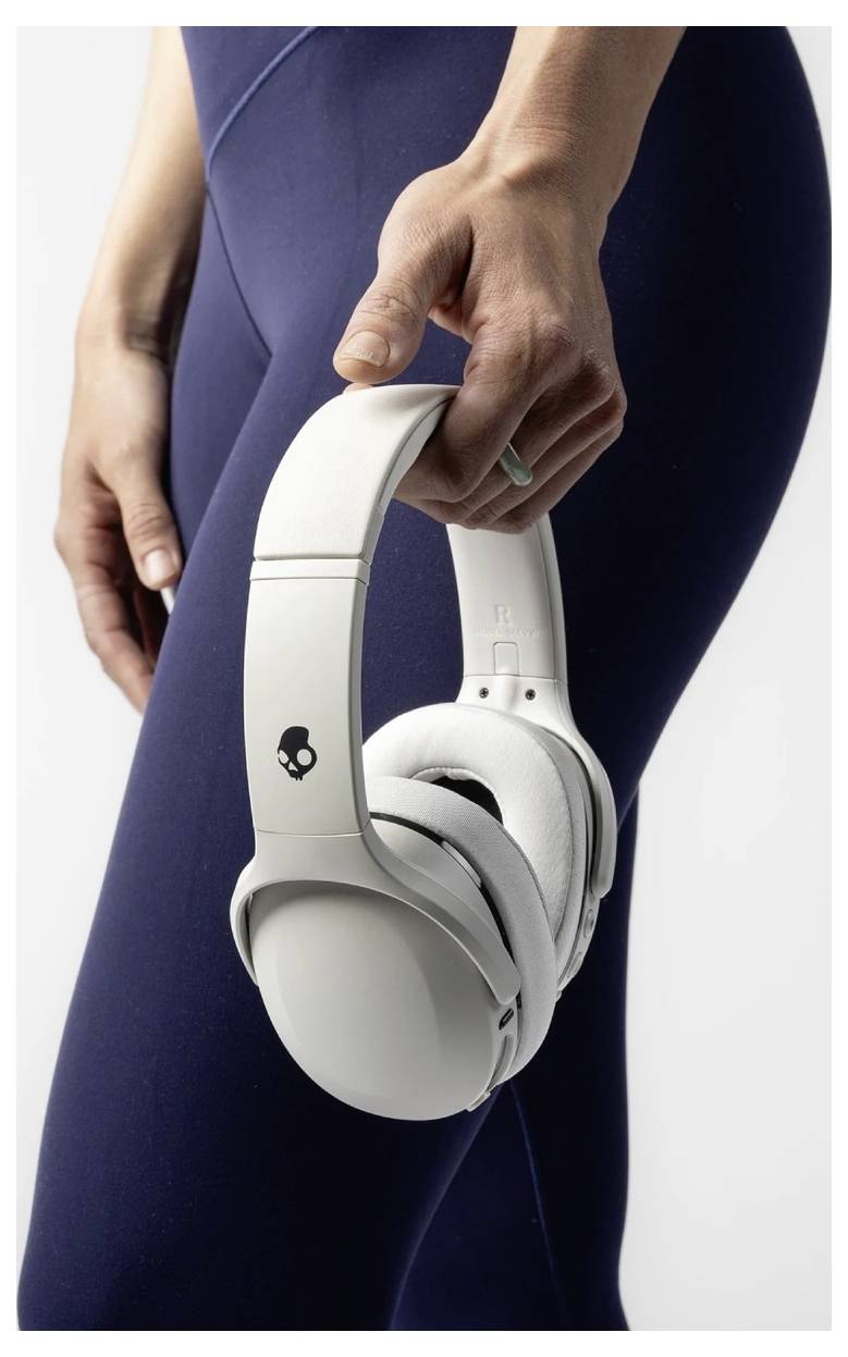 Eine Person in dunkelblauen Leggings hält weiße Over-Ear-Kopfhörer mit einem kleinen Totenkopf-Logo auf der Ohrmuschel gegen einen einfachen Hintergrund.