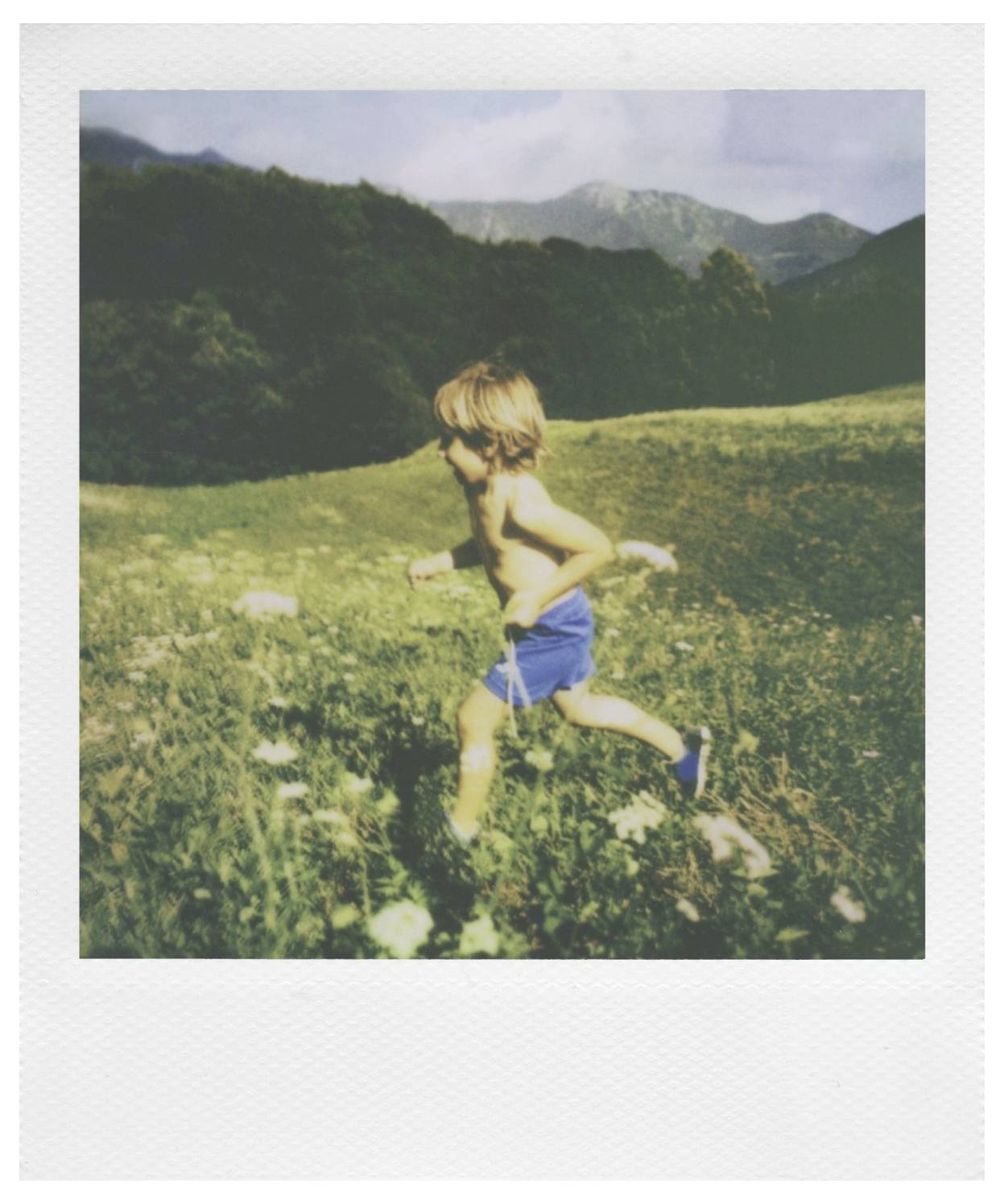 Ein Kind in blauen Shorts läuft durch ein Grasfeld mit Bergen im Hintergrund an einem sonnigen Tag.