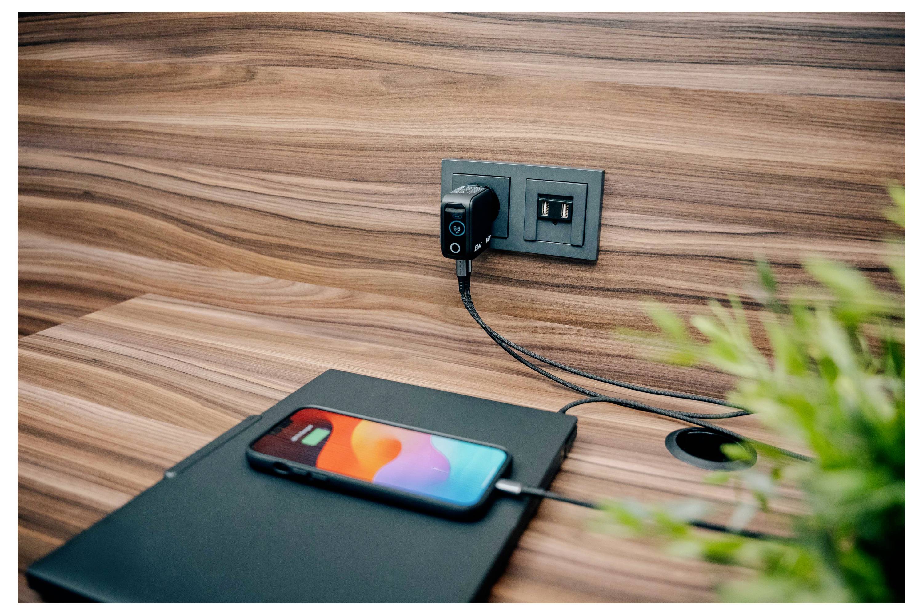 Ein Smartphone lädt auf einem Holzschreibtisch auf und ist in eine schwarze Wandsteckdose eingesteckt. Ein Topfpflanze und ein Notizbuch sind in der Nähe.