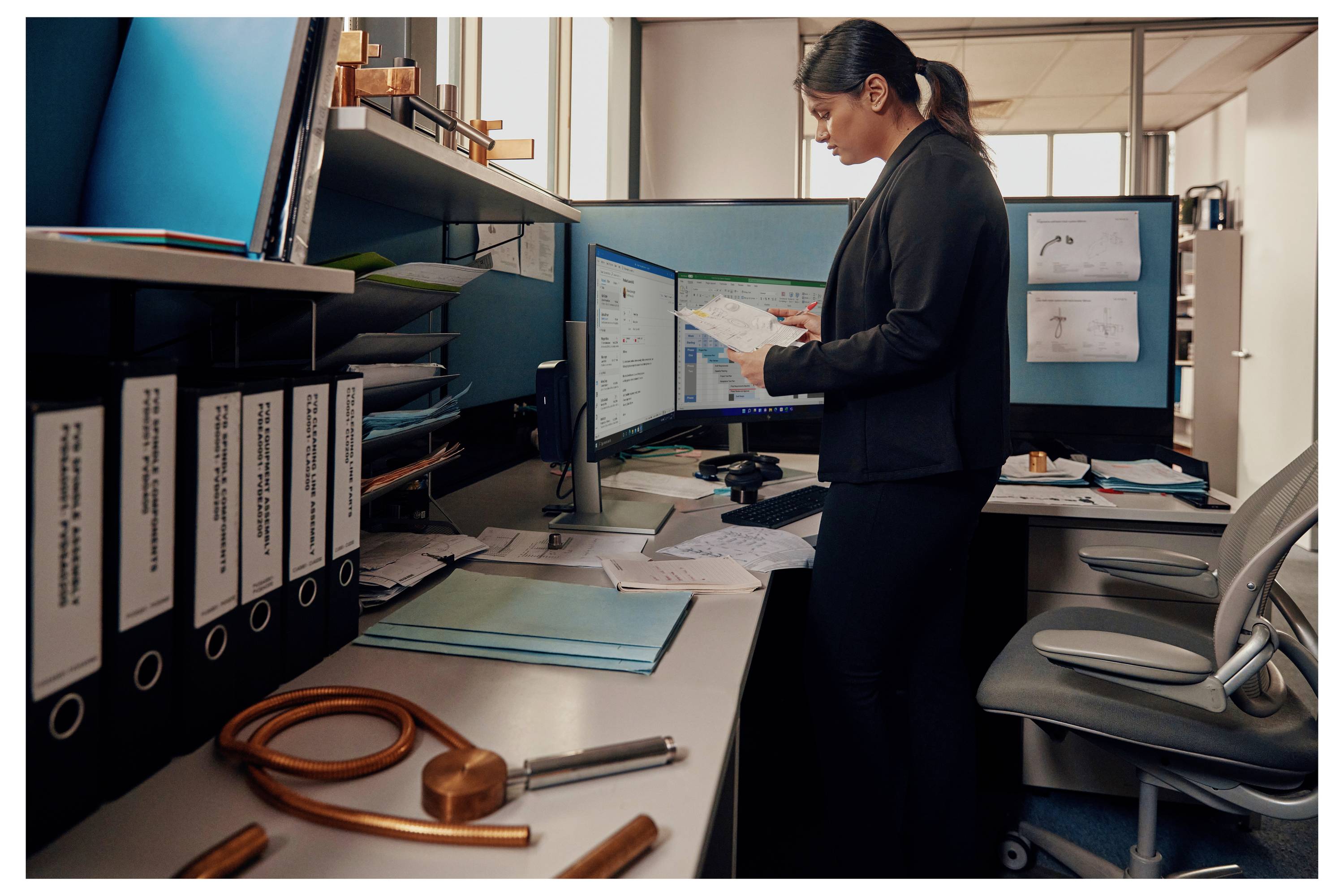 Eine Person in einem Büro steht an einem Schreibtisch und überprüft Dokumente auf zwei Monitoren. In der Nähe befinden sich Regale mit Akten und ein Stethoskop.