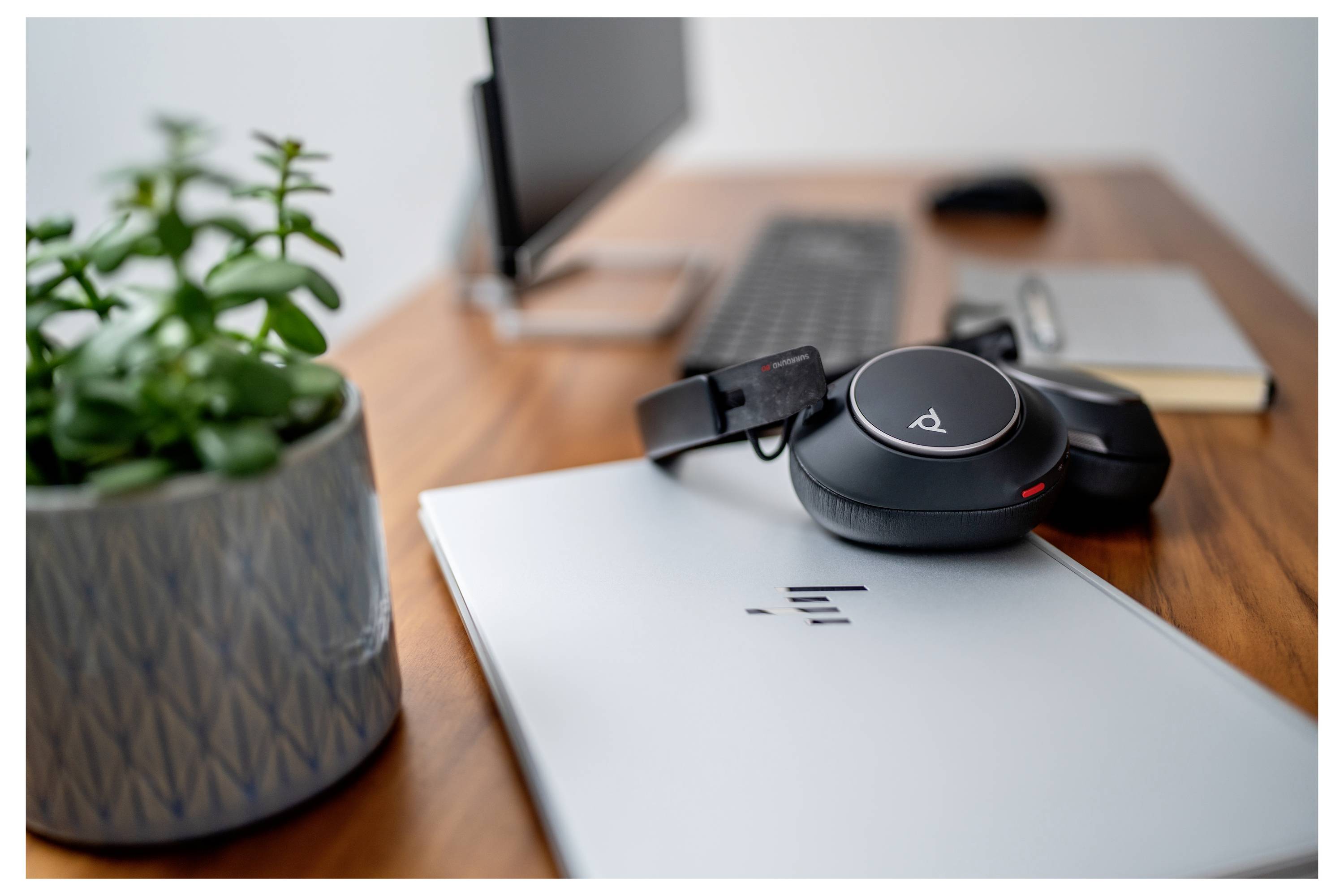 Ein eleganter Arbeitsplatz mit einem Laptop, kabellosen Kopfhörern, einer kleinen Pflanze, einem Desktop-Monitor, einer Tastatur und einem geschlossenen Notizbuch auf einem Holzschreibtisch.