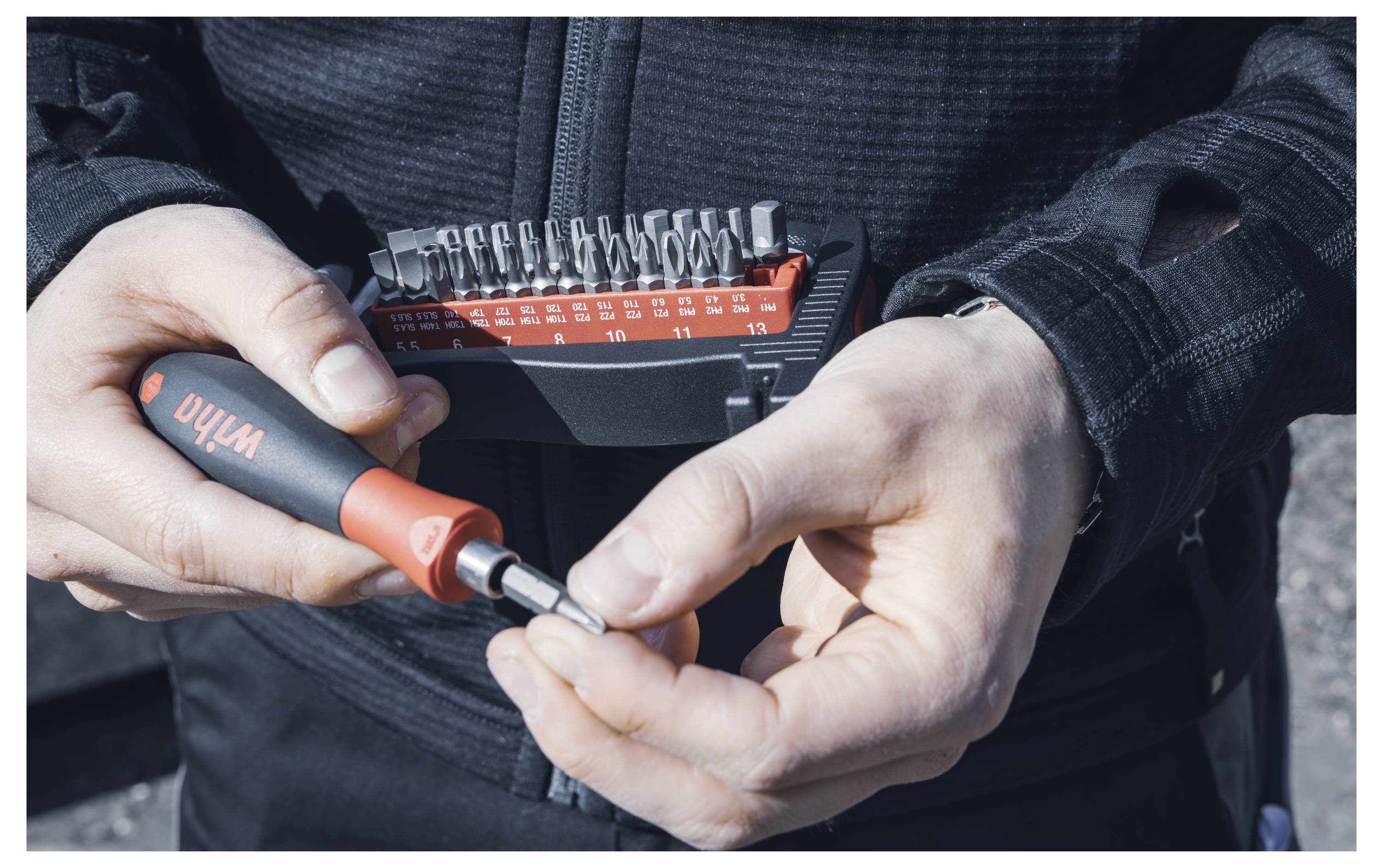 Eine Person in dunkler Kleidung hält einen rot-schwarzen Werkzeugsatz mit austauschbaren Bits und bereitet sich darauf vor, einen Bit am Griff anzubringen.