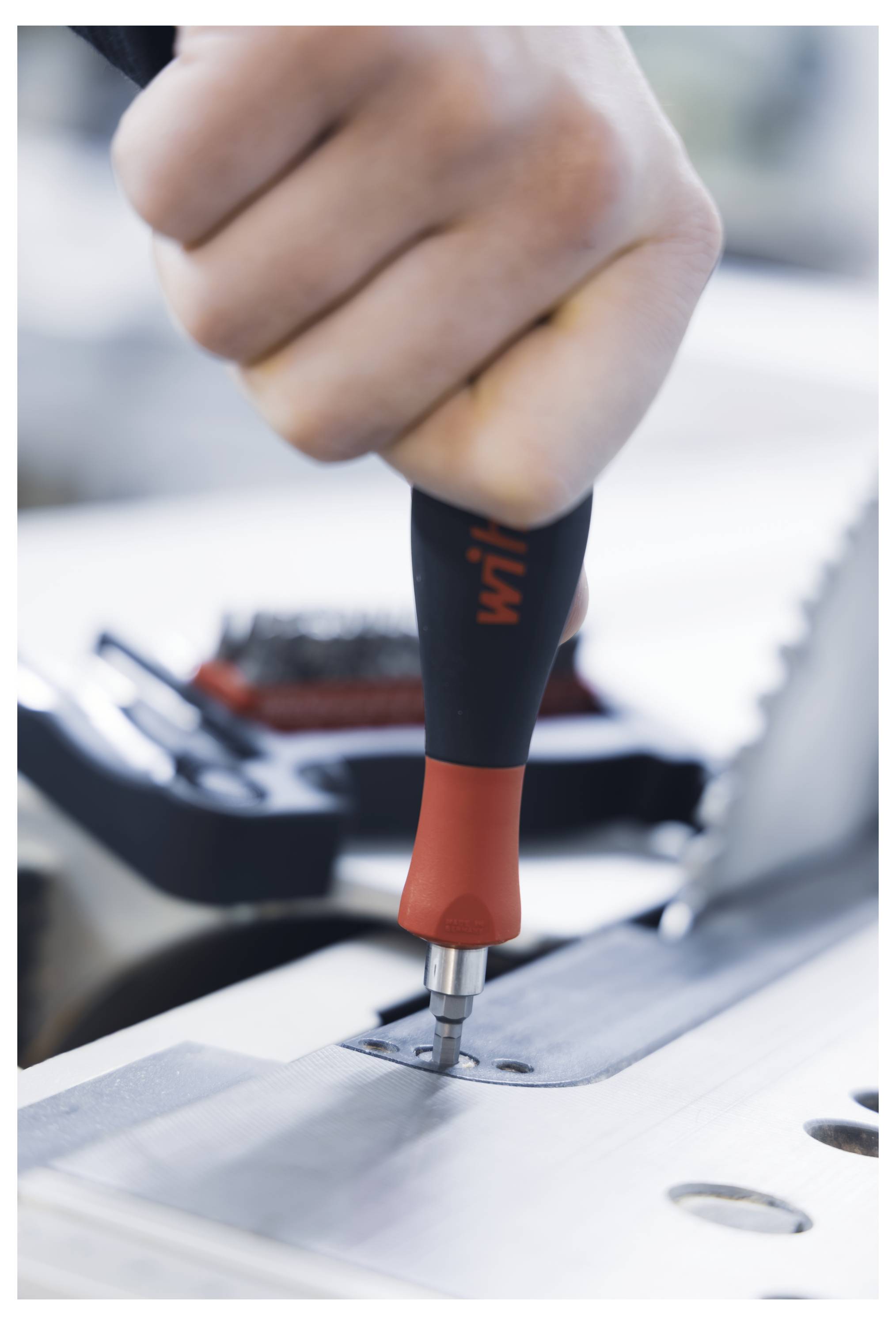 Eine Hand verwendet einen roten und schwarzen Schraubendreher, um eine Schraube auf einer Metalloberfläche festzuziehen, mit einem unscharfen Sägeblatt im Hintergrund.