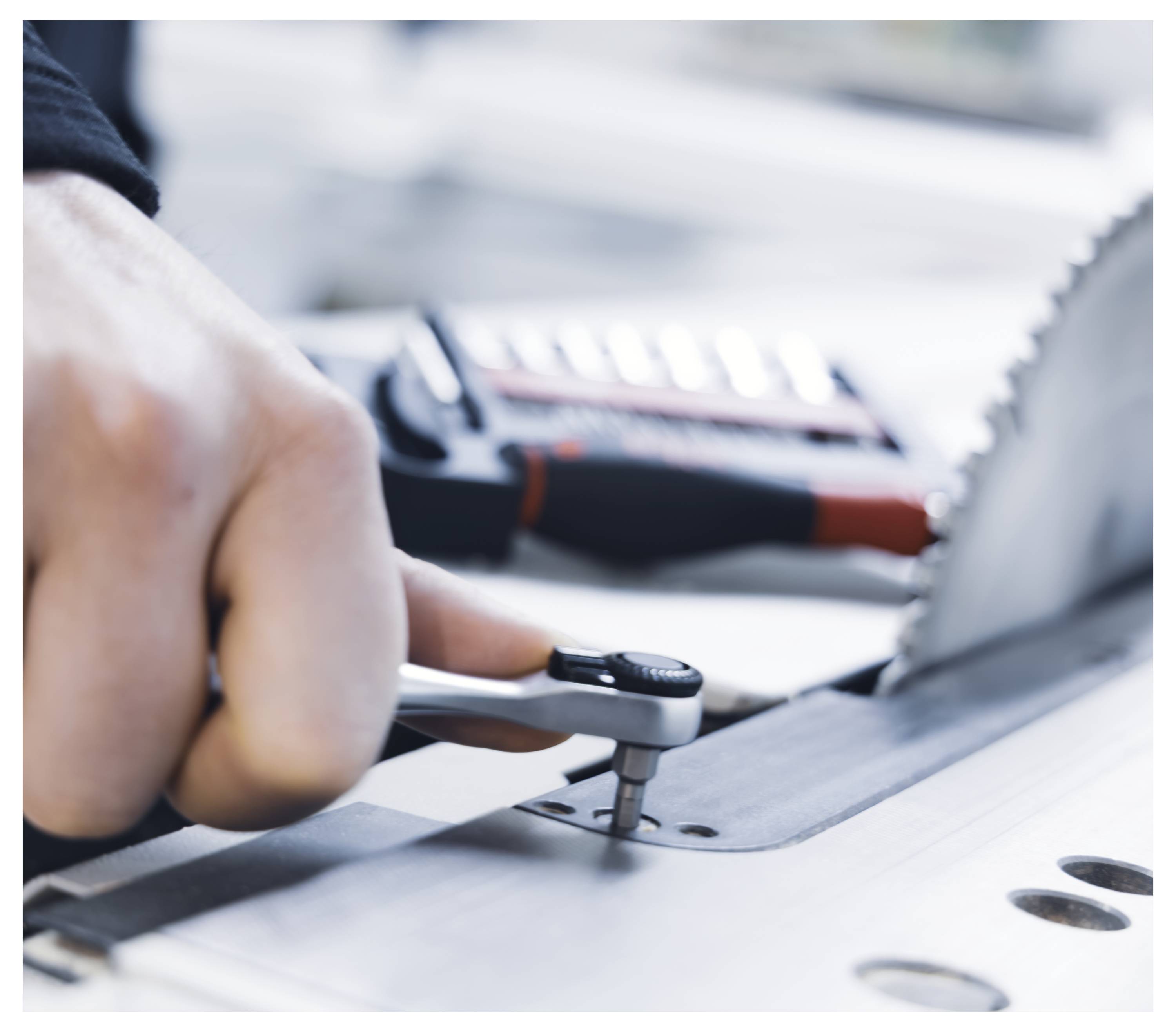 Eine Hand benutzt einen Schraubenschlüssel, um einen Bolzen neben einer rotierenden Sägeblatt anzupassen, mit einer unscharfen Werkzeugkiste im Hintergrund.