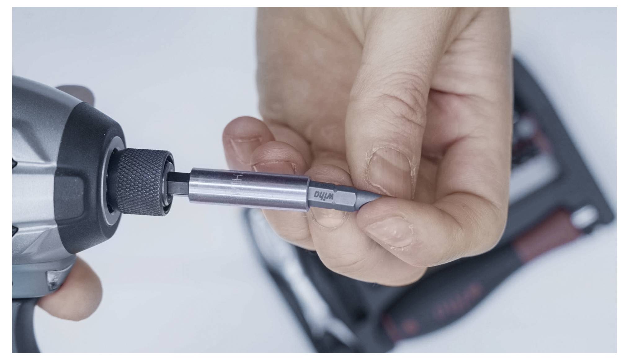 Eine Hand führt einen Bohrer in einen elektrischen Schraubendreher ein, mit unscharfen Werkzeugen im Hintergrund.