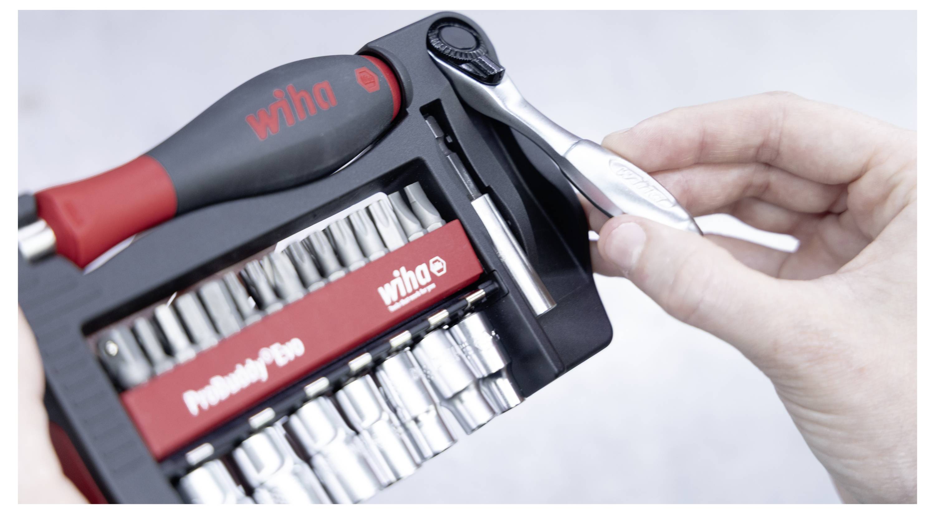 Eine Hand, die einen kleinen Ratschenschlüssel über einem Werkzeugkasten mit verschiedenen Schraubendreher-Bits und Stecknüssen hält, mit dem Wiha-Branding.