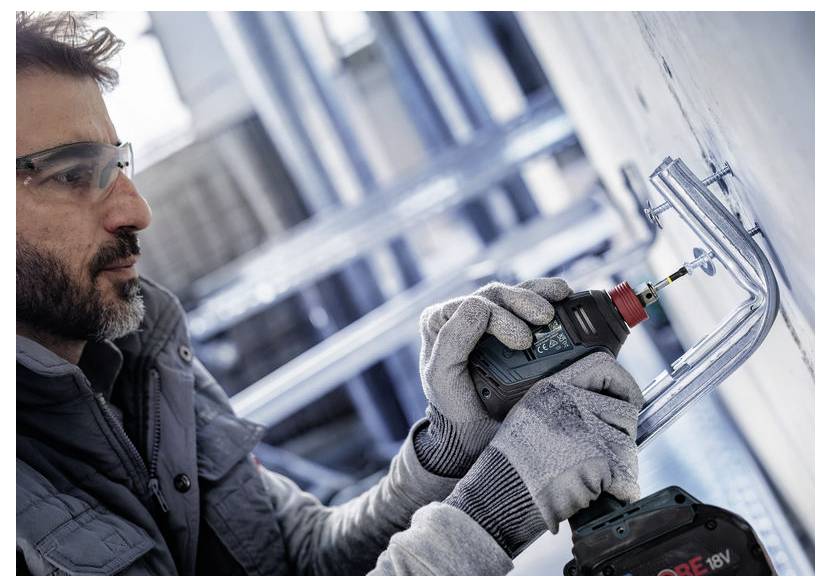Eine Person mit Schutzbrille und Handschuhen verwendet einen Elektroschrauber, um Metallhalterungen an einer Wand in einer Industrieumgebung zu befestigen.