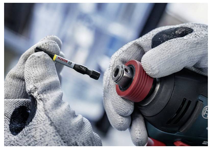 Eine Person mit Handschuhen hält einen Elektrobohrer und einen Bohraufsatz mit einem „Bosch"-Etikett und bereitet sich vor, den Aufsatz am Bohrer anzubringen.