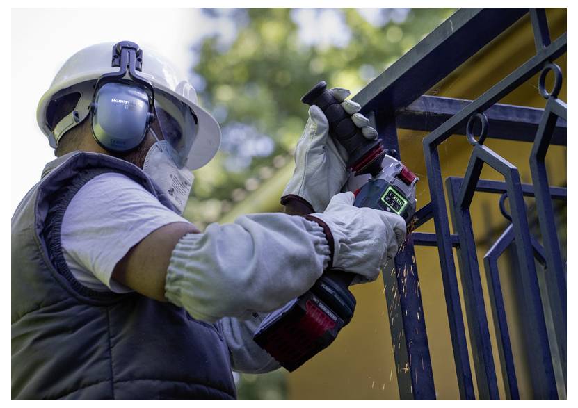 Ein Arbeiter, der Schutzausrüstung trägt, einschließlich Handschuhen, einem Helm und Gehörschutz, verwendet ein Elektrowerkzeug zum Schneiden von Metall an einem Geländer im Freien.