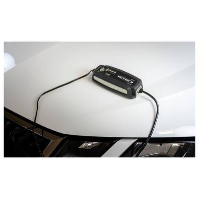Ein schwarzes Batterieladegerät sitzt auf der Motorhaube eines weißen Fahrzeugs mit angeschlossenen Kabeln, was darauf hindeutet, dass es die Batterie des Fahrzeugs auflädt.