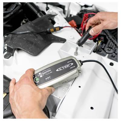 Eine Person verbindet ein CTEK-Ladegerät mit einer Autobatterie, wobei rote und schwarze Kabel sichtbar sind. Die Motorhaube ist geöffnet.