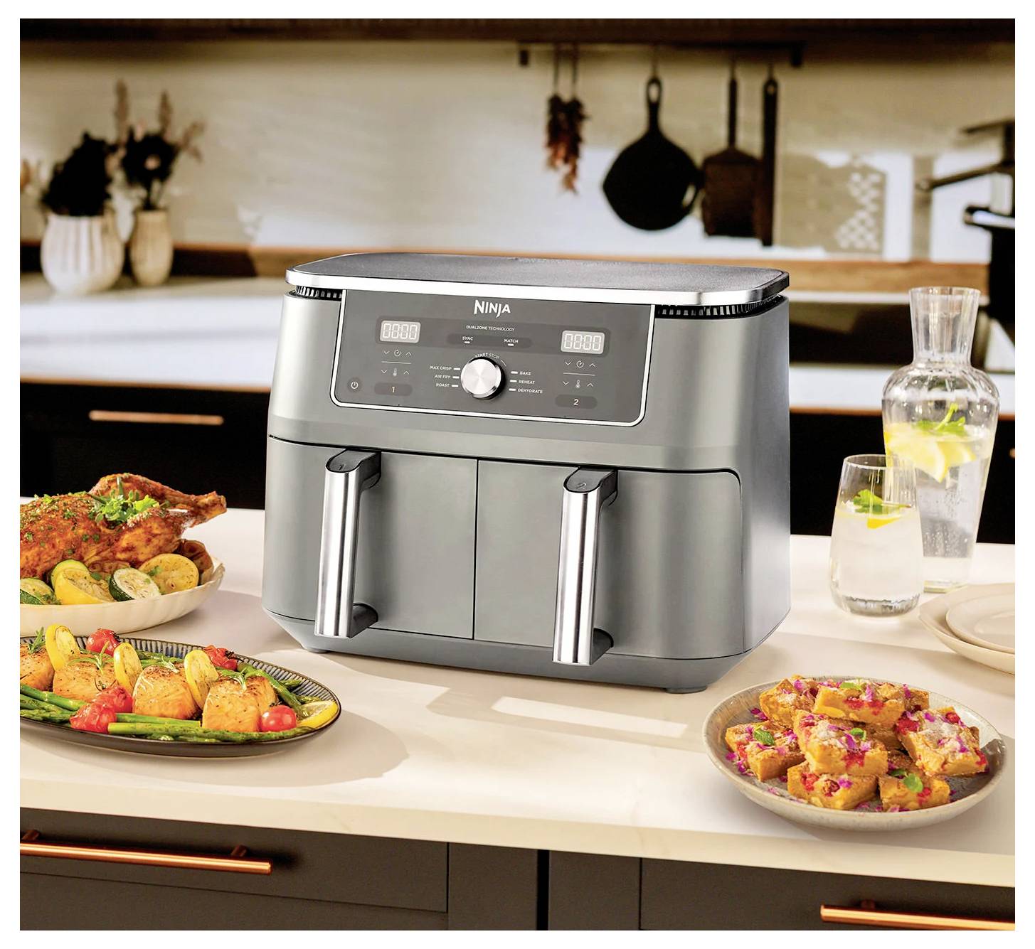 Grauer Heißluftfritteusen-Gerät auf der Küchentheke mit gegartem Gemüse, Hähnchen und Snacks auf Tellern rundherum. Moderne Küche im Hintergrund.