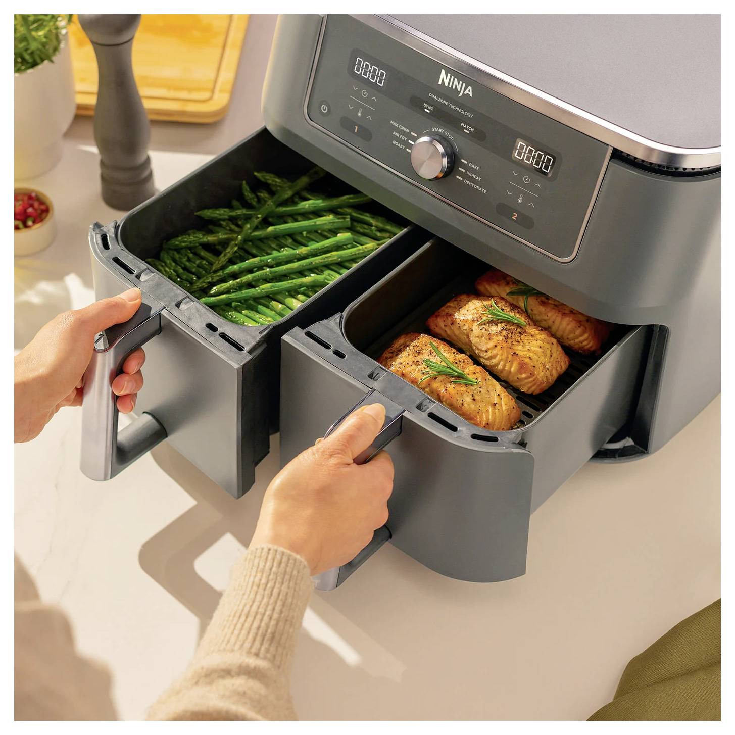 Eine Person bereitet Speisen in einer Heißluftfritteuse vor und zeigt Spargel in einem Fach und Lachs in einem anderen, beide gegart und servierfertig.