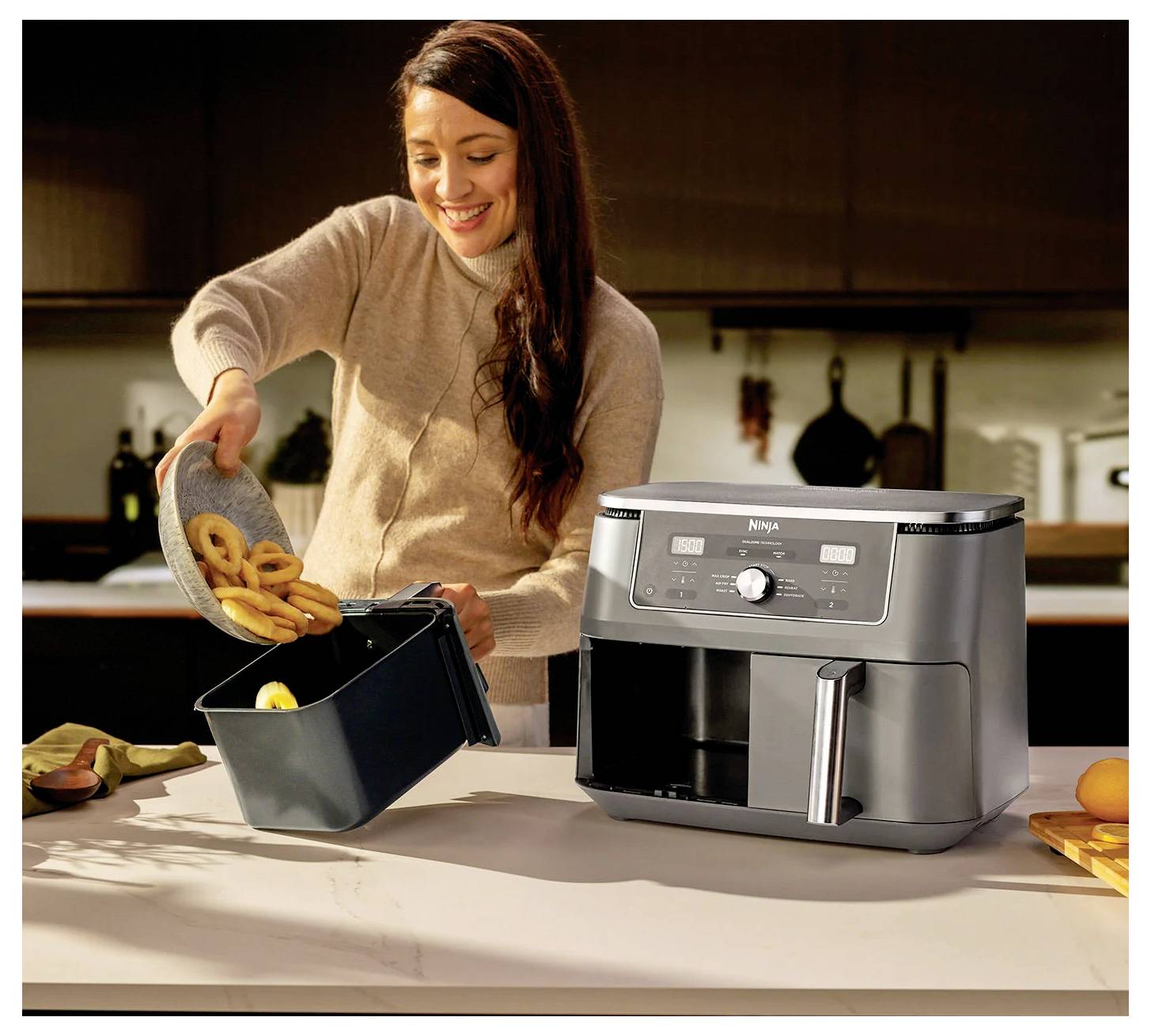 Eine Frau lächelt, während sie frittierte Ringe in einen Korb einer Heißluftfritteuse in einer Küche legt, mit Küchenutensilien und Orangen im Hintergrund.