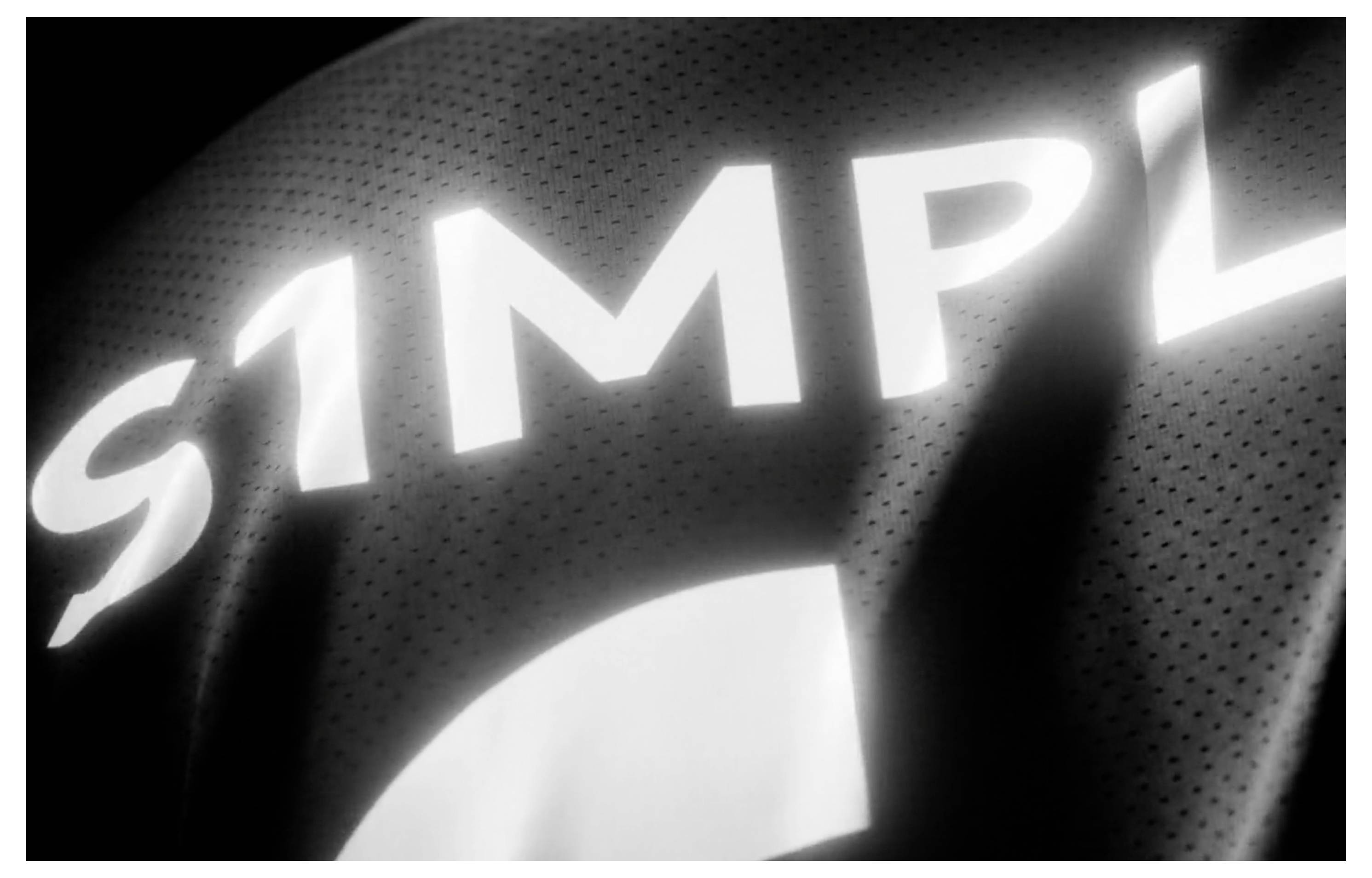 Nahaufnahme eines schwarzen Sporttrikots mit großen weißen Buchstaben „SIMP" und einem weißen gebogenen Logo, dargestellt in Graustufen.