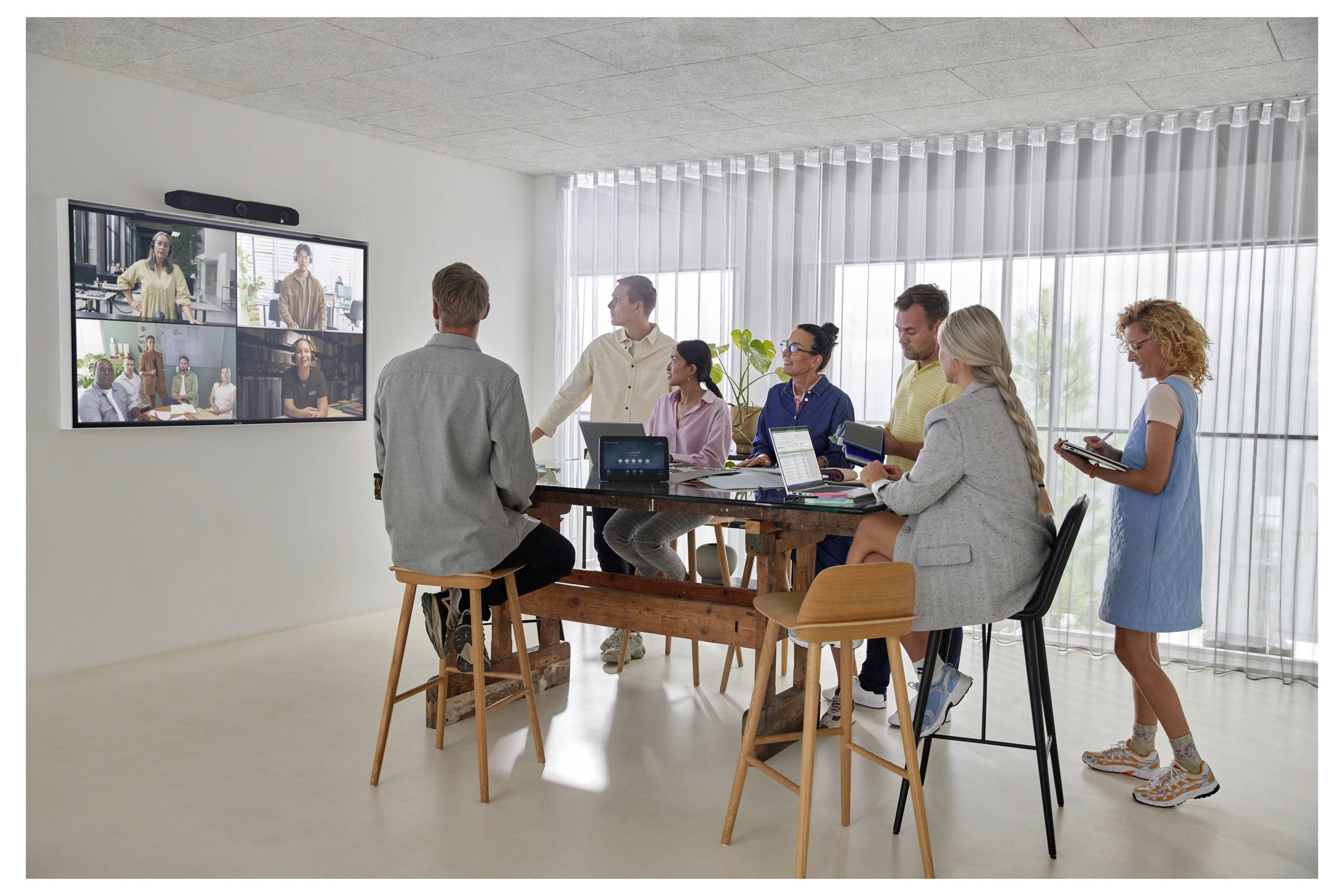 Eine kleine Gruppe in einem modernen Besprechungszimmer beobachtet eine Videokonferenz mit vier Personen auf einem an der Wand montierten Bildschirm, während sie um einen hohen Tisch mit Laptops und Notizbüchern stehen.