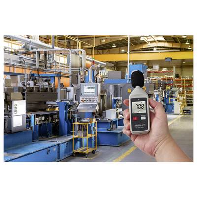 Hand hält ein digitales Schallpegelmessgerät, das 70,8 dB in einer großen Industriefabrik mit Maschinen und Rohren anzeigt.