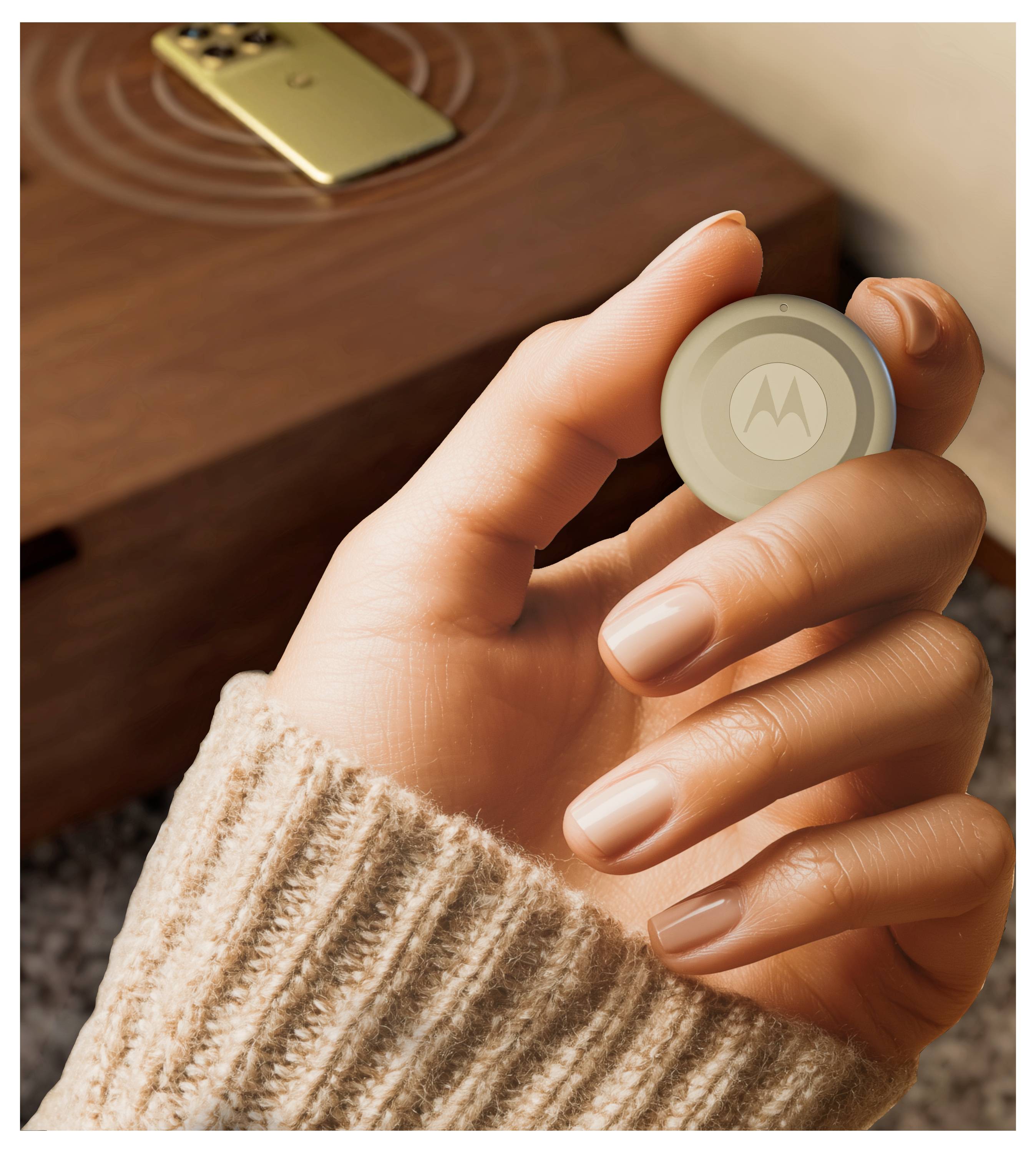 Eine Hand hält ein kleines, rundes, hellfarbiges Gerät mit dem Motorola-Logo. Ein Smartphone liegt auf einer Holzoberfläche im Hintergrund.