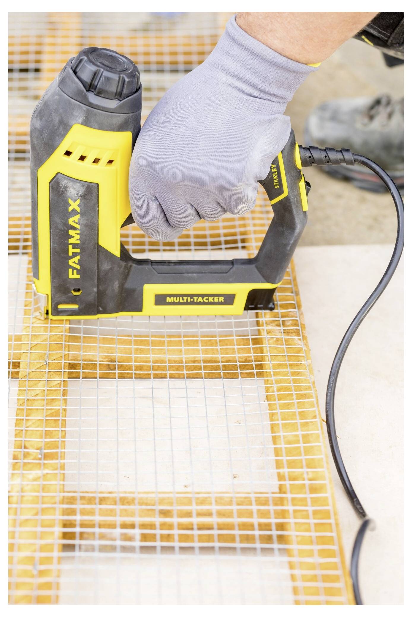 Eine Person trägt Handschuhe und benutzt einen gelb-schwarz gefärbten elektrischen Hefter mit der Bezeichnung "Fatmax Multi-Tacker" auf einem Netzstoffmaterial über Holz.
