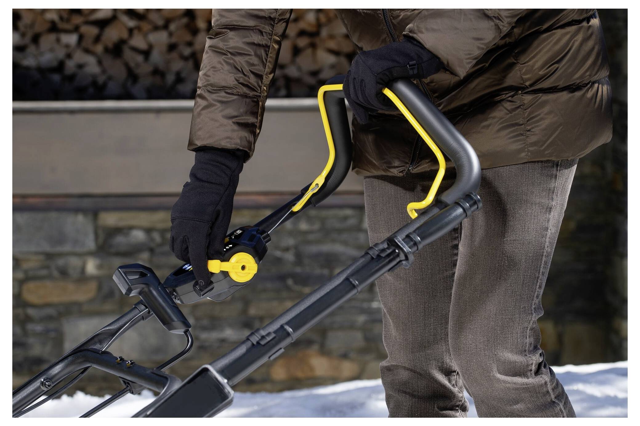 Eine Person in einem braunen Mantel und schwarzen Handschuhen stellt den Griff eines Schneefräers auf einer verschneiten Oberfläche ein, mit einer Steinmauer im Hintergrund.