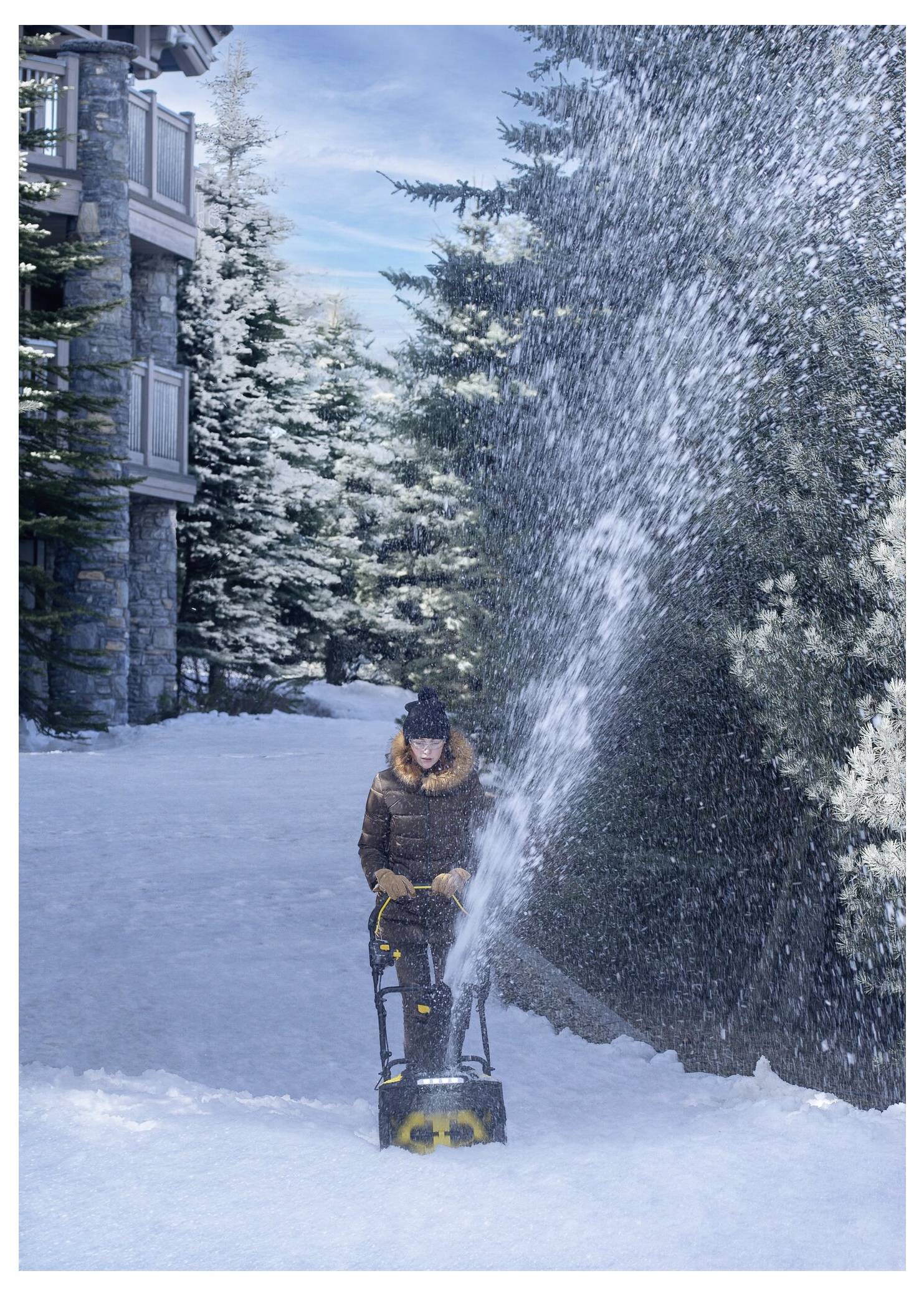 Eine Person nutzt einen Schneefräser, um einen Weg in einer verschneiten Landschaft neben einem Steingebäude und immergrünen Bäumen freizumachen.