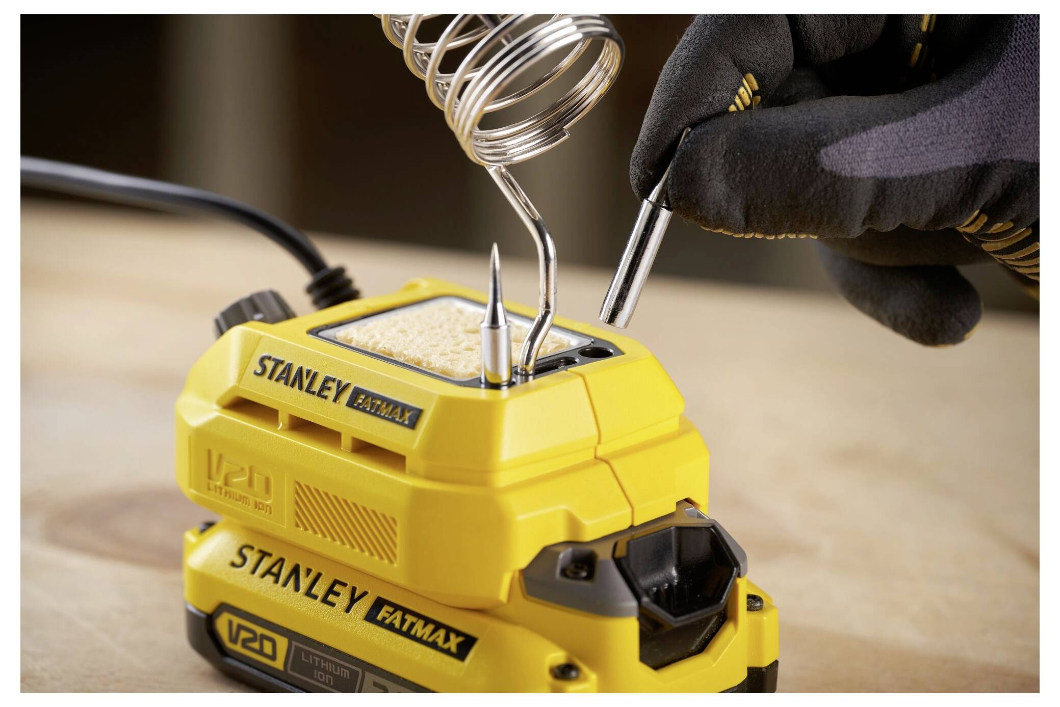 Eine Person in Handschuhen bedient ein Stanley FatMax V20 Lötgerät auf einer Holzoberfläche, wobei ein Lötkolben auf seinem Halter ruht.