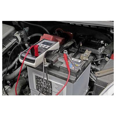 Ein digitales Multimeter mit roten und schwarzen Messspitzen prüft eine Autobatterie im Motorraum.