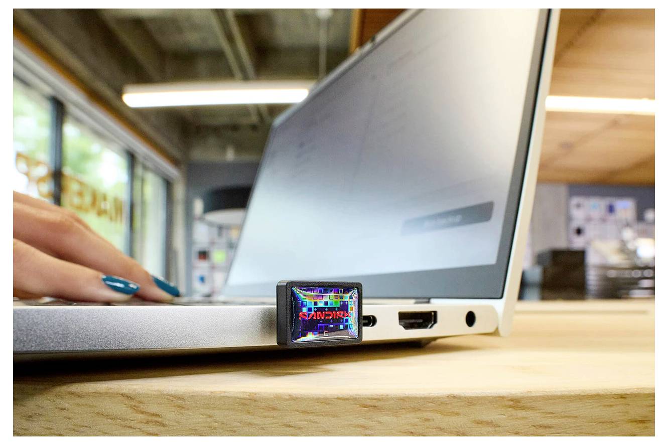 Eine Hand führt ein kleines, farbiges elektronisches Gerät in einen USB-Port eines Laptops ein, in einem hellen Raum mit großen Fenstern und Holzoberflächen.